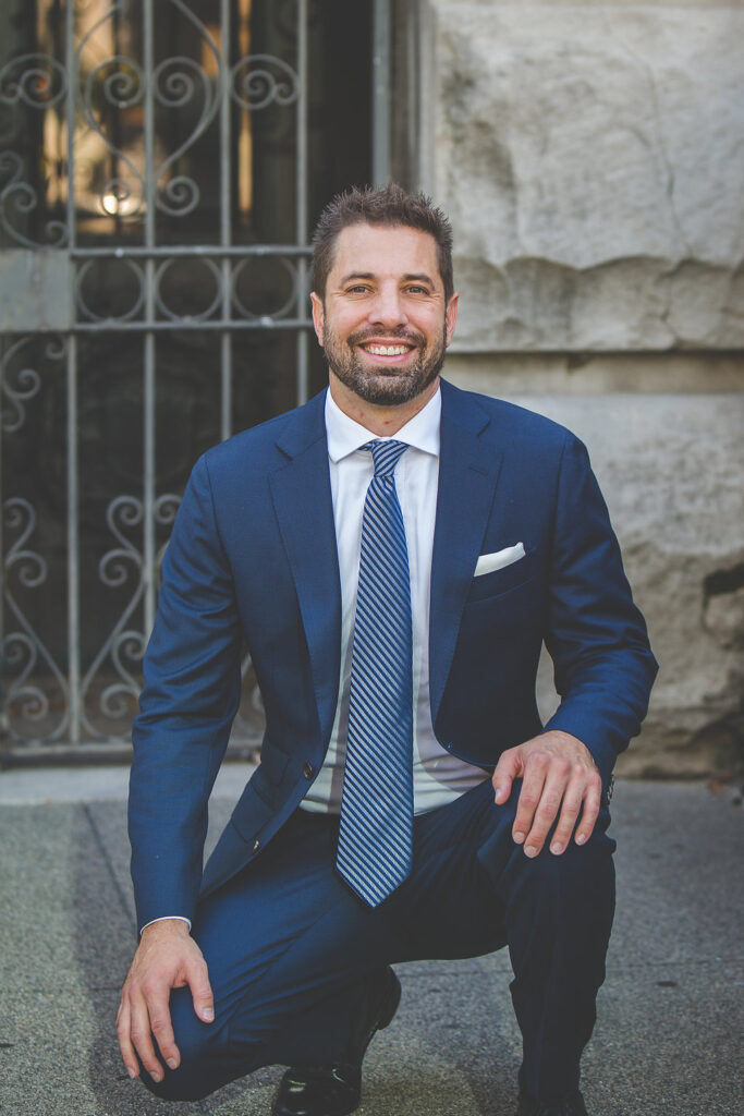 Joshua Wilson of Joshua Wilson Consulting. Navy blue suit, blue tie, white shirt. Josh is on one knee and smiling in front of Monument Circle in downtown, Indianpolis.